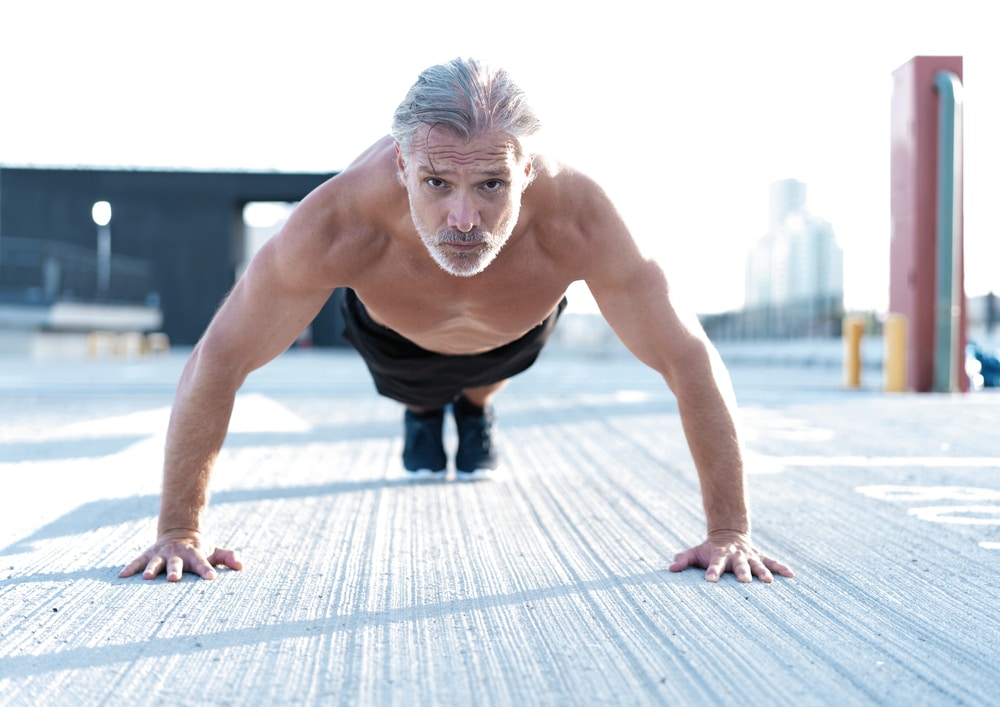 Middle aged man does pushups on concrete while looking directly into the camera